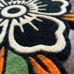 Macro texture shot of the Inked Garden Rug, emphasizing the hand-tufted wool pile of the front pattern and the clean fabric backing with neat red stitching along the detailed scalloped edge.