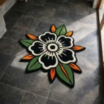 Perspective view of the rectangular Inked Garden Rug, featuring a traditional tattoo flash-style repeating pattern of red roses and stars on a cream background with a scalloped black border, styled on a modern concrete floor next to a textured white pouf and a mid-century wooden cabinet bathed in natural sunlight.