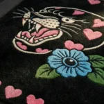 Angled close-up view of the Inkheart Panther rug, featuring a hand-tufted traditional tattoo panther head, a bright blue flower, and pink hearts against a solid black background.