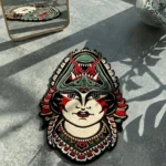 Top-down flat view of the die-cut Lady Face traditional tattoo rug, featuring a woman wearing a fierce green and red demon headdress, styled on a smooth concrete floor with striped sunlight shadows.