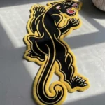 Angled perspective view of the black Neo-Traditional Panther rug styled on a warm rustic hardwood floor in a cozy den, positioned near a classic brown leather armchair and a wooden coffee table.