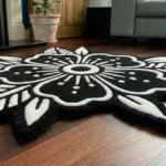 Angled view of the Obsidian Bloom black and white floral shaped rug resting on dark hardwood floors in a cozy room, positioned near a lit stone fireplace hearth and the leg of a brown leather armchair.