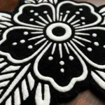 Low-angle close-up view of the Obsidian Bloom rug on a hardwood floor, showcasing the plush thickness of the hand-tufted wool and the deep carving that defines the white traditional tattoo flower design against the black base.