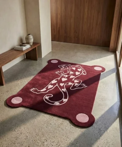 Perspective view of the burgundy Panther of Hearts rug styled in a sunlit, modern entryway on a smooth light concrete floor, placed near a warm wooden bench and doorway.