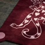 Close-up detail of the burgundy Panther of Hearts rug on a dark tile floor, showcasing the plush thickness of the hand-tufted wool, the solid pink circular corner accent, and the expertly carved pink panther tail and claws.