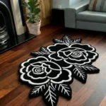 Wide perspective view of the Petal Duet black and white double rose rug resting on dark hardwood floors in a warm den, surrounded by a wooden bookcase, a glowing floor lamp, a lit stone fireplace, and a brown leather armchair.