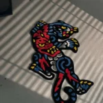 Top-down flat view of the die-cut Phantom Panther rug resting on a smooth light floor, featuring a vibrant red, blue, yellow, and black abstract traditional tattoo design, beautifully bathed in sharp, striped sunlight shadows.