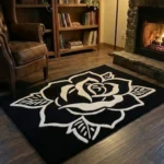 Perspective view of the Phantom Rose black rectangular rug with a white traditional tattoo rose design, styled on a dark hardwood floor in a warm den featuring a lit stone fireplace, a wooden bookcase, and a brown leather armchair.