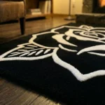 Low-angle close-up view of the Phantom Rose rug, showcasing the plush thickness of the hand-tufted black wool and the crisp 3D carved white rose petals, with a lit stone fireplace in the blurred background.