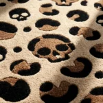 Close-up macro detail of the Savage Skull Leopard rug bathed in sunlight, highlighting the dense hand-tufted wool pile and the crisp 3D carving of a brown skull motif within the leopard print pattern.