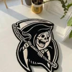 Angled perspective view of the Shadow Reaper black and white traditional tattoo flash rug resting on a white wooden floor, bathed in bright natural sunlight.