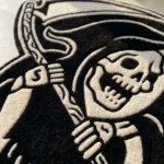 Close-up macro detail of the Shadow Reaper rug in sunlight, highlighting the dense hand-tufted black wool pile and the crisp 3D carved white lines defining the grim reaper's skull face and scythe.