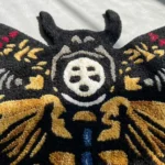 Close-up macro detail of the Skull Death Moth rug bathed in natural light, highlighting the totally dense texture of the hand-tufted wool and the precise 3D carving around the central white skull face and gold wing joints.