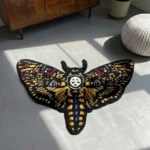 Elevated perspective view of the entire Skull Death Moth tattoo flash rug on a grey concrete floor, beautifully showcasing its full wingspan, colorful gold, red, and blue wing spots, and striking central skull motif next to a white pouf.