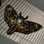 High-angle perspective view of the die-cut Skull Death Moth traditional tattoo rug resting on a smooth grey concrete floor, featuring a black base with intricate gold wings and a central white skull, bathed in striped sunlight shadows.