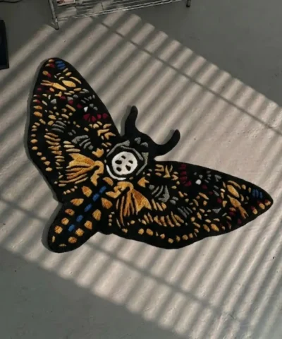 High-angle perspective view of the die-cut Skull Death Moth traditional tattoo rug resting on a smooth grey concrete floor, featuring a black base with intricate gold wings and a central white skull, bathed in striped sunlight shadows.