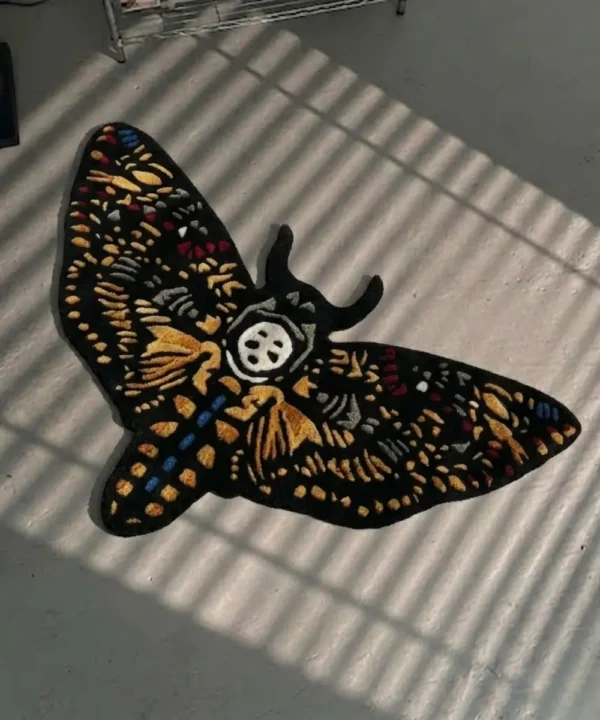 High-angle perspective view of the die-cut Skull Death Moth traditional tattoo rug resting on a smooth grey concrete floor, featuring a black base with intricate gold wings and a central white skull, bathed in striped sunlight shadows.