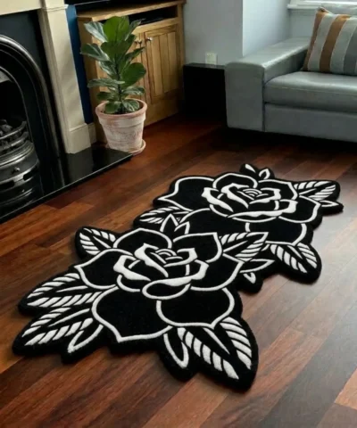 Perspective view of the Twin Ink Rose black and white double flower tattoo rug styled on a dark wood floor in a living room next to a potted plant, a black fireplace, and a light grey sofa.