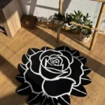 Perspective view of the Void Blossom black and white rose-shaped rug styled on a light hardwood floor in a sunlit room, next to a potted plant and a wooden ladder shelf.