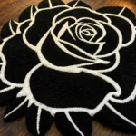 Angled close-up view of the Void Blossom rose rug on a dark wood floor, highlighting the dense black hand-tufted wool pile and the thick, crisp white carved lines defining the petals.