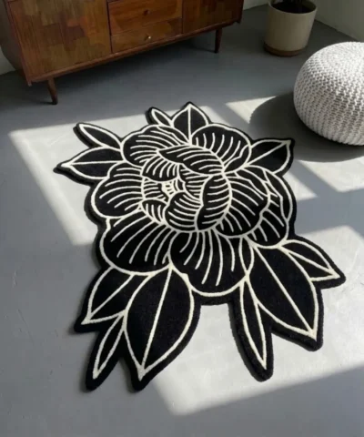 A full top-down studio shot of the Flash Flower rug, featuring an American Traditional tattoo-inspired floral motif in high-contrast black and white, styled on a wooden floor near a vibrant orange speaker and plant.