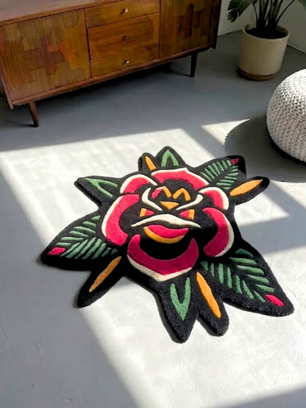 Perspective view of the rectangular American Traditional Rose Rug, featuring a traditional tattoo flash-style repeating pattern of two red roses and stars on a cream background with a scalloped black border, styled on a modern concrete floor next to a textured white pouf and a mid-century wooden cabinet bathed in natural sunlight.