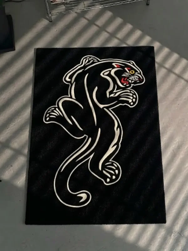 Top-down flat view of the die-cut Black Panther Tattoo Rug, resting on a grey concrete floor, showcasing its irregular shape, crouching panther silhouette, and vibrant red, yellow, and blue traditional tattoo-style face and border accents.