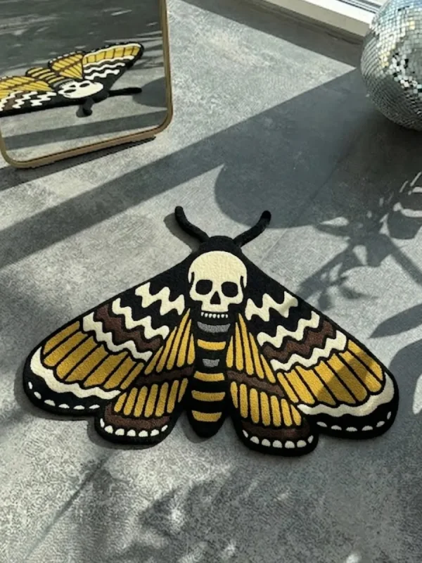 Top-down view of the hand-tufted Death Head Moth rug, in traditional tattoo style with gold, black, and cream colors, lying flat on a grey concrete floor with natural sunlight shadows.