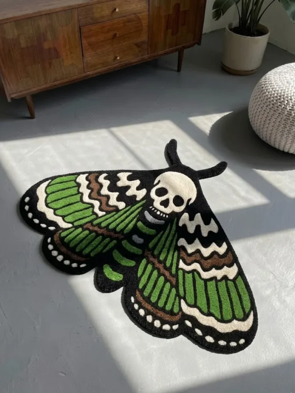 High-angle view of the Death Moth neo-traditional tattoo flash rug resting on a smooth grey concrete floor next to a white pouf, with dramatic window shadows cast across its skull and wing details.