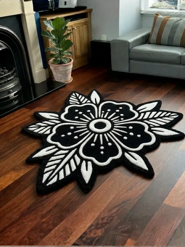 Perspective view of the Obsidian Bloom black and white traditional tattoo flower rug styled on a wooden floor in a living room next to a classic black fireplace, a potted plant, and a light grey sofa.