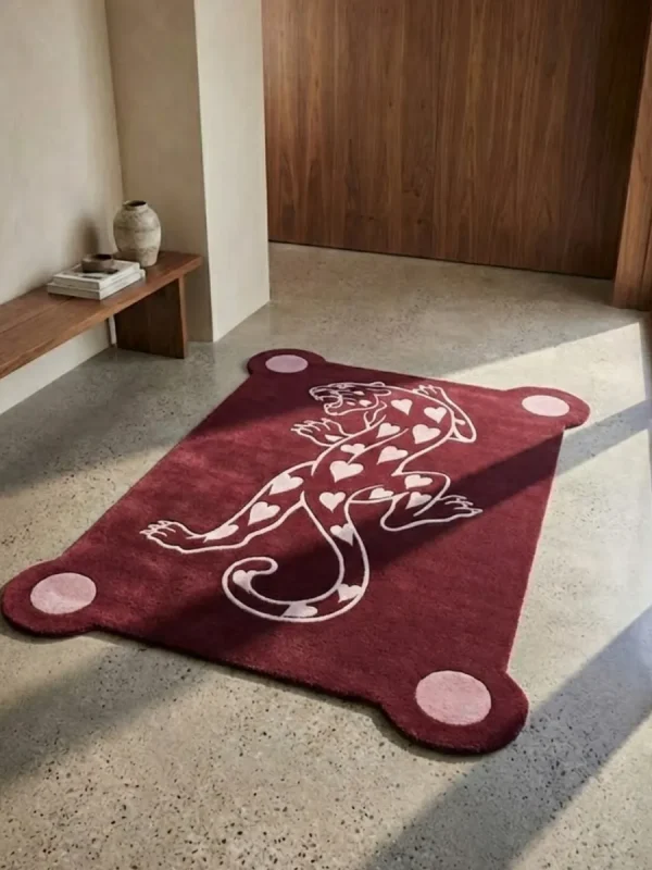 Perspective view of the burgundy Panther of Hearts rug styled in a sunlit, modern entryway on a smooth light concrete floor, placed near a warm wooden bench and doorway.