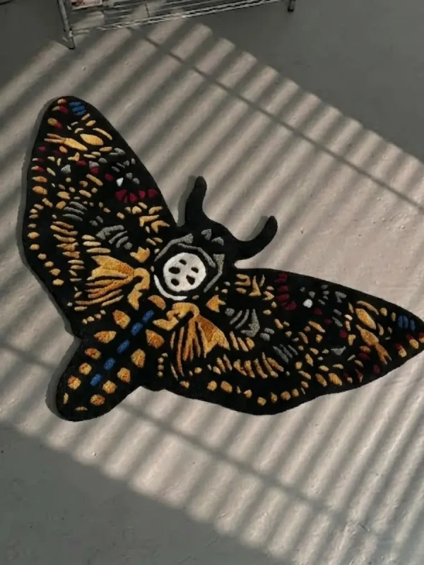 High-angle perspective view of the die-cut Skull Death Moth traditional tattoo rug resting on a smooth grey concrete floor, featuring a black base with intricate gold wings and a central white skull, bathed in striped sunlight shadows.